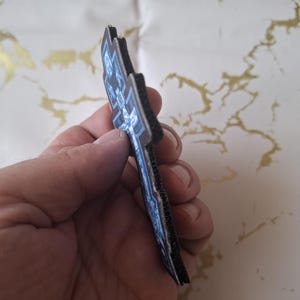 Pu&ograve; includere: Una toppa blu scuro e nera con un design dettagliato. La toppa ha una superficie strutturata e un bordo nero. Il design sembra essere una croce o un emblema stilizzato. La toppa &egrave; tenuta in una mano.