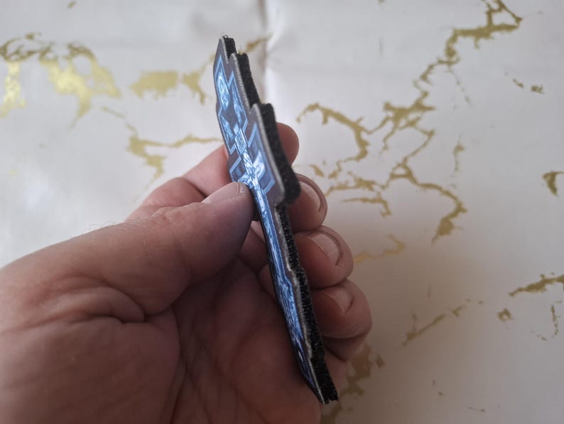 Pu&ograve; includere: Una toppa blu scuro e nera con un design dettagliato. La toppa ha una superficie strutturata e un bordo nero. Il design sembra essere una croce o un emblema stilizzato. La toppa &egrave; tenuta in una mano.