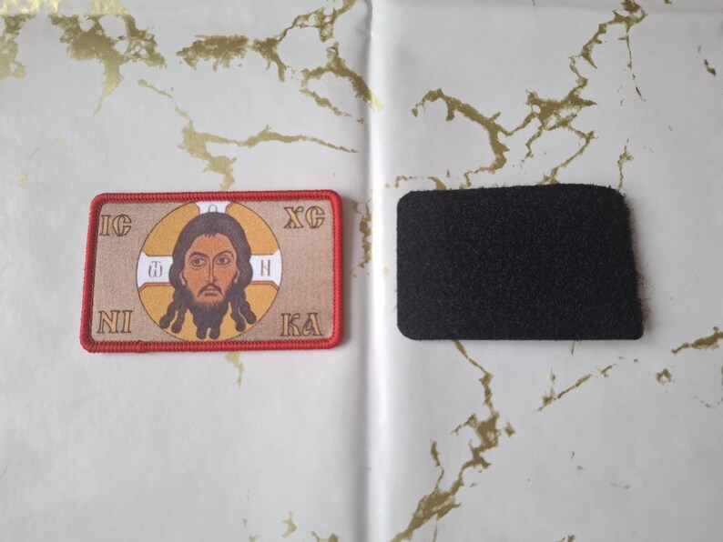 Peut inclure: Un &eacute;cusson rectangulaire avec une ic&ocirc;ne religieuse, fond marron, bordure rouge et texte. &Agrave; c&ocirc;t&eacute;, un patch de fixation auto-agrippante noir et rectangulaire. L'ic&ocirc;ne repr&eacute;sente un homme avec une barbe et des cheveux longs.