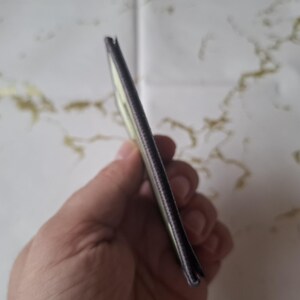 May include: A slim, rectangular wallet with a dark exterior and a lighter interior is held in a hand. The wallet appears to be made of a textured material, possibly leather or a similar fabric. The edges are neatly finished.