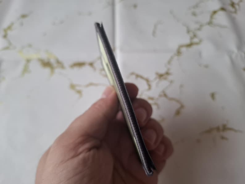 May include: A slim, rectangular wallet with a dark exterior and a lighter interior is held in a hand. The wallet appears to be made of a textured material, possibly leather or a similar fabric. The edges are neatly finished.