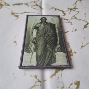 May include: Rectangular patch featuring a religious icon of an angel with a sword and shield. The angel is depicted in a flowing robe, with wings and a halo. The patch has a gray border and is set against a white and gold background.
