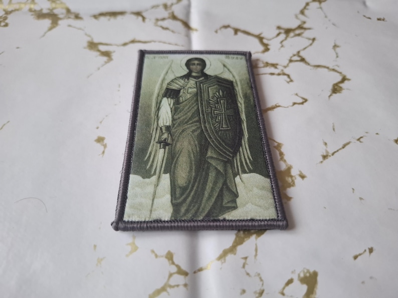 May include: Rectangular patch featuring a religious icon of an angel with a sword and shield. The angel is depicted in a flowing robe, with wings and a halo. The patch has a gray border and is set against a white and gold background.