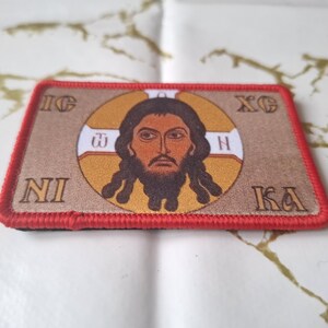 Peut inclure: Un &eacute;cusson rectangulaire avec une bordure rouge pr&eacute;sente une image centrale d'un homme avec une barbe et de longs cheveux, entour&eacute; d'un halo dor&eacute;. Le fond est beige et l'&eacute;cusson contient du texte en grec.