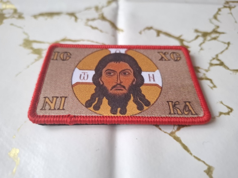 Peut inclure: Un &eacute;cusson rectangulaire avec une bordure rouge pr&eacute;sente une image centrale d'un homme avec une barbe et de longs cheveux, entour&eacute; d'un halo dor&eacute;. Le fond est beige et l'&eacute;cusson contient du texte en grec.