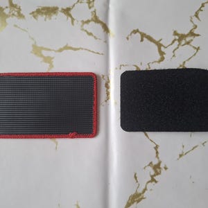 Peut inclure: Deux patchs rectangulaires. L'un est noir avec un bord rouge et une surface textur&eacute;e. L'autre est un patch noir uni avec une texture floue. Les deux patchs sont sur une surface blanche avec des marbrures dor&eacute;es.