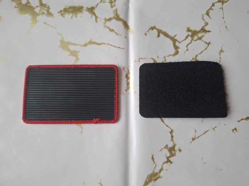 Peut inclure: Deux patchs rectangulaires. L'un est noir avec un bord rouge et une surface textur&eacute;e. L'autre est un patch noir uni avec une texture floue. Les deux patchs sont sur une surface blanche avec des marbrures dor&eacute;es.