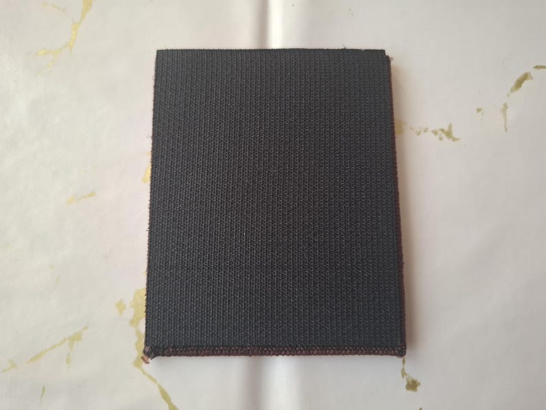 K&ouml;nnte beinhalten: Ein quadratischer, dunkelgrauer Stofffleck mit strukturierter Oberfl&auml;che. Die Kanten sind mit einem d&uuml;nnen, dunkelroten Rand versehen. Der Aufn&auml;her befindet sich auf einem wei&szlig;-gold gemusterten Hintergrund.