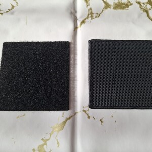 May include: Two black square patches, one with a textured, fuzzy surface and the other with a woven, grid-like pattern. The patches are displayed on a white surface with gold-colored marbling.