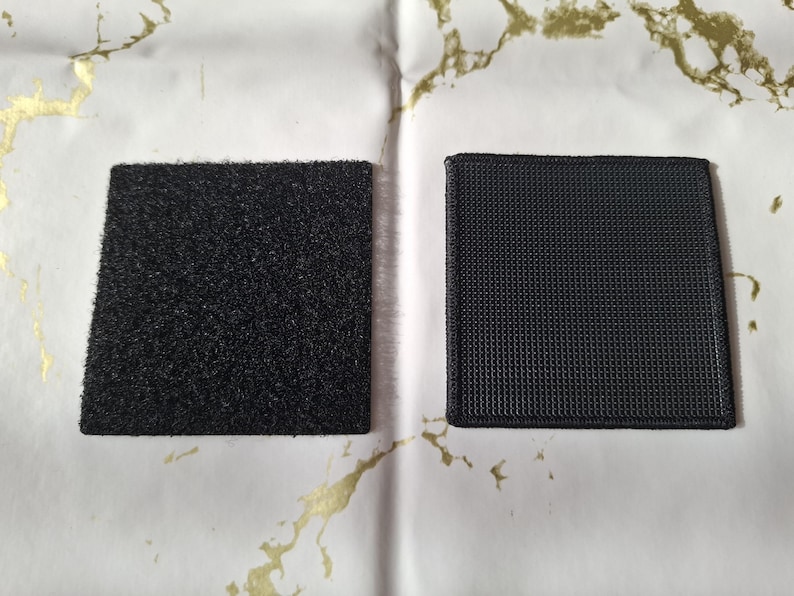 May include: Two black square patches, one with a textured, fuzzy surface and the other with a woven, grid-like pattern. The patches are displayed on a white surface with gold-colored marbling.