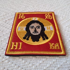 Op de afbeelding: Geborduurde patch met een religieus icoon. De patch heeft een rode achtergrond met gouden randen en letters. Een centrale gouden cirkel bevat een afbeelding van een gezicht met donker haar. De patch is vierkant met een licht trapeziumvormige vorm.