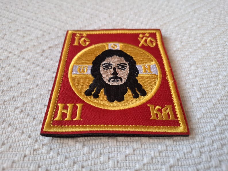 Op de afbeelding: Geborduurde patch met een religieus icoon. De patch heeft een rode achtergrond met gouden randen en letters. Een centrale gouden cirkel bevat een afbeelding van een gezicht met donker haar. De patch is vierkant met een licht trapeziumvormige vorm.
