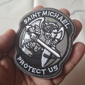 May include: A circular embroidered patch depicting Saint Michael with a sword and shield. The patch is predominantly grey and white, with the text "SAINT MICHAEL PROTECT US" in white lettering.