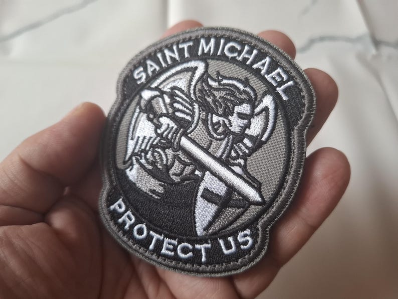 May include: A circular embroidered patch depicting Saint Michael with a sword and shield. The patch is predominantly grey and white, with the text "SAINT MICHAEL PROTECT US" in white lettering.