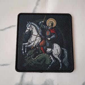 Pode incluir: Um remendo bordado preto com um cavalo branco e um cavaleiro de armadura montado nele. O cavaleiro está segurando uma lança e usando uma capa vermelha. O cavalo está em pé sobre um dragão verde.