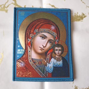 Könnte beinhalten: Ein rechteckiger Aufnäher mit einer religiösen Ikone einer Frau und eines Kindes. Die Frau trägt eine rote Kopfbedeckung und hält das Kind, das blau gekleidet ist. Das Bild hat einen goldenen Heiligenschein und wird von einem blauen Rand umrahmt.