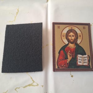 K&ouml;nnte beinhalten: Eine Ikone von Jesus Christus, mit Heiligenschein und Buch, wird neben einem dunkelgrauen, rechteckigen Filzst&uuml;ck gezeigt. Die Ikone hat einen braunen Rahmen und ist vor einem hellen Hintergrund platziert.