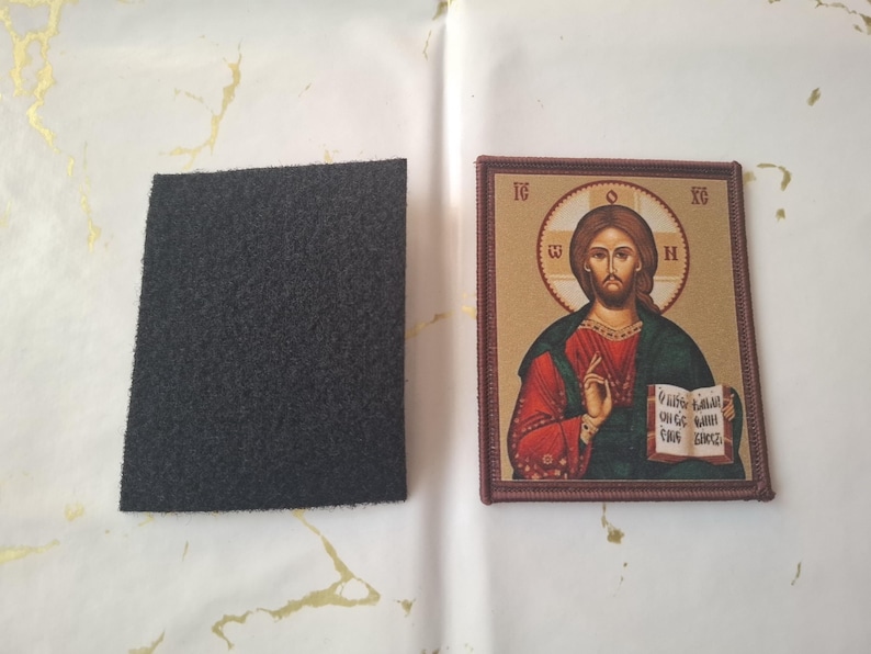 K&ouml;nnte beinhalten: Eine Ikone von Jesus Christus, mit Heiligenschein und Buch, wird neben einem dunkelgrauen, rechteckigen Filzst&uuml;ck gezeigt. Die Ikone hat einen braunen Rahmen und ist vor einem hellen Hintergrund platziert.