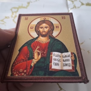 K&ouml;nnte beinhalten: Rechteckige religi&ouml;se Ikone mit einer Darstellung von Jesus Christus. Er wird mit Heiligenschein gezeigt und h&auml;lt ein Buch mit Text. Die Ikone hat einen braunen Rand und ist haupts&auml;chlich in Rot-, Gr&uuml;n- und Goldt&ouml;nen gehalten.