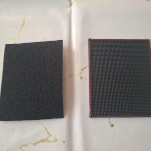 K&ouml;nnte beinhalten: Zwei rechteckige Aufn&auml;her. Einer ist schwarz, mit einer flauschigen, strukturierten Oberfl&auml;che. Der andere ist dunkelgrau mit einem Gittermuster und einem rotbraunen Rand. Beide werden auf einer wei&szlig;en Oberfl&auml;che mit goldenen Akzenten angezeigt.