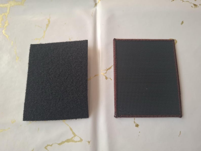 K&ouml;nnte beinhalten: Zwei rechteckige Aufn&auml;her. Einer ist schwarz, mit einer flauschigen, strukturierten Oberfl&auml;che. Der andere ist dunkelgrau mit einem Gittermuster und einem rotbraunen Rand. Beide werden auf einer wei&szlig;en Oberfl&auml;che mit goldenen Akzenten angezeigt.