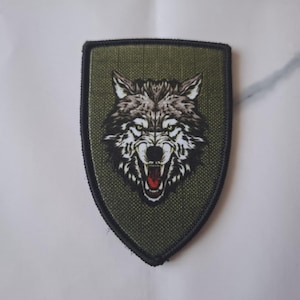 Peut inclure: Écusson en forme de bouclier avec une tête de loup détaillée. Le loup est gris et blanc, avec une langue rouge et des yeux verts, sur un fond vert. L'écusson a une bordure noire et est probablement destiné aux vêtements ou accessoires.