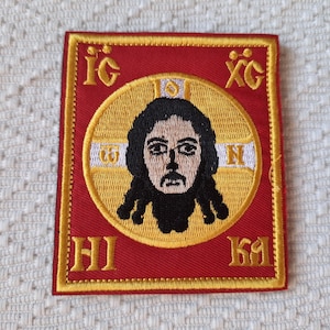 Könnte beinhalten: Gestickter Aufnäher mit einem religiösen Symbol. Der quadratische Aufnäher hat einen roten Hintergrund und einen goldenen Rand. Das zentrale Bild ist eine Darstellung einer Figur mit dunklem Haar und Bart, umgeben von einem goldenen Heiligenschein.