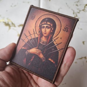 May include: A rectangular patch depicting a religious icon of a woman with a halo, holding swords. The image has a warm colour palette with brown and gold tones. The patch has a brown border.