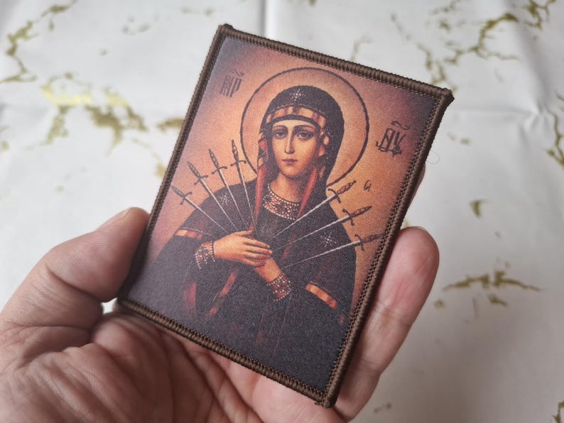May include: A rectangular patch depicting a religious icon of a woman with a halo, holding swords. The image has a warm colour palette with brown and gold tones. The patch has a brown border.