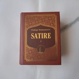 Può includere: Un libro con copertina rigida marrone con dettagli dorati. Il titolo "SATIRE" è in lettere dorate, con il nome dell'autore, Radoje Domanović, sopra. La copertina presenta un bordo dorato decorativo. Un'opera letteraria.