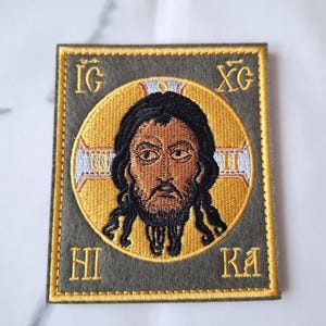 Peut inclure: Écusson brodé représentant une icône religieuse. L'image centrale est celle d'un homme aux cheveux noirs et à la barbe, entouré d'un halo doré. L'écusson a une bordure dorée et un fond vert olive avec des lettres grecques.