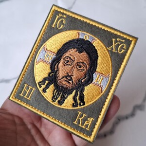 Op de afbeelding: Geborduurde patch met een religieus icoon. De patch heeft een rechthoekige vorm met een gouden rand. De centrale afbeelding is een figuur met donker haar en een baard, omgeven door een gouden halo. De achtergrond is olijfgroen.