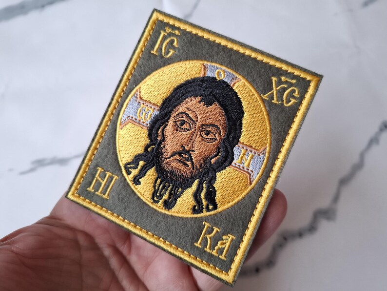 Op de afbeelding: Geborduurde patch met een religieus icoon. De patch heeft een rechthoekige vorm met een gouden rand. De centrale afbeelding is een figuur met donker haar en een baard, omgeven door een gouden halo. De achtergrond is olijfgroen.