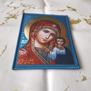 K&ouml;nnte beinhalten: Ein rechteckiger Aufn&auml;her mit einem religi&ouml;sen Bild einer Frau und eines Kindes, umrahmt von einem blauen Rand. Die Ikone hat einen goldenen Heiligenschein und ist in warmen Farben mit roten, blauen und goldenen Akzenten gehalten.