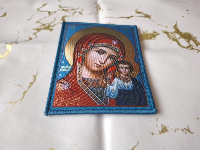 K&ouml;nnte beinhalten: Ein rechteckiger Aufn&auml;her mit einem religi&ouml;sen Bild einer Frau und eines Kindes, umrahmt von einem blauen Rand. Die Ikone hat einen goldenen Heiligenschein und ist in warmen Farben mit roten, blauen und goldenen Akzenten gehalten.