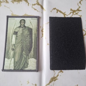 May include: An embroidered patch featuring an image of an angel holding a shield and sword. The patch has a rectangular shape with a purple border and a black hook and loop fastener backing.