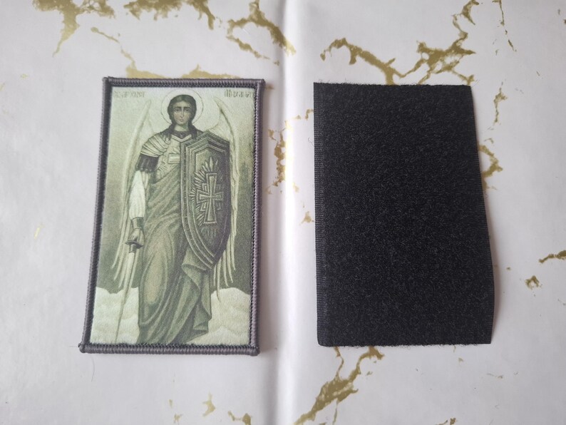 May include: An embroidered patch featuring an image of an angel holding a shield and sword. The patch has a rectangular shape with a purple border and a black hook and loop fastener backing.