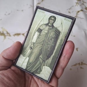 May include: Rectangular patch featuring an illustration of an angel with a shield and sword. The angel is depicted in shades of green, white, and gray, with a halo above its head. The patch has a gray border and is held in a hand.