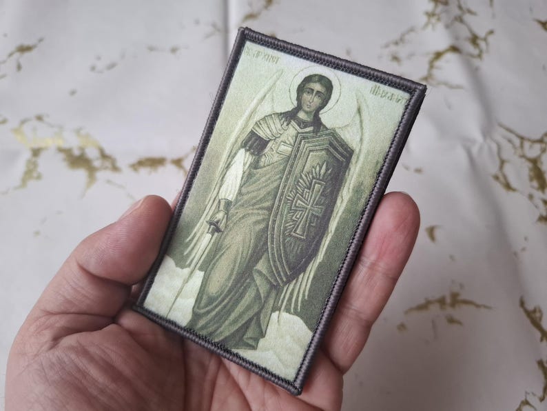 May include: Rectangular patch featuring an illustration of an angel with a shield and sword. The angel is depicted in shades of green, white, and gray, with a halo above its head. The patch has a gray border and is held in a hand.