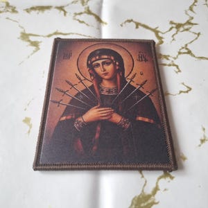 May include: A rectangular religious icon depicting a woman with a halo, hands clasped, and seven swords piercing her. It has a brown border, set against a white and gold background. The image is a religious item.