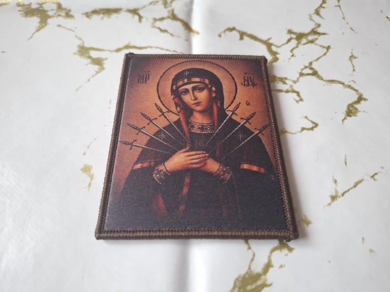 May include: A rectangular religious icon depicting a woman with a halo, hands clasped, and seven swords piercing her. It has a brown border, set against a white and gold background. The image is a religious item.