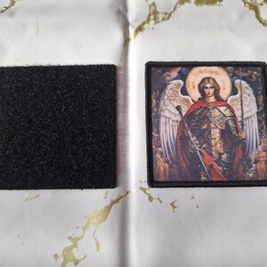May include: A square black hook and loop fastener patch next to a rectangular embroidered patch. The embroidered patch features a detailed image of an angel with a halo, wings, and a sword, in shades of red, gold, and brown.