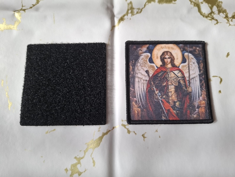 May include: A square black hook and loop fastener patch next to a rectangular embroidered patch. The embroidered patch features a detailed image of an angel with a halo, wings, and a sword, in shades of red, gold, and brown.