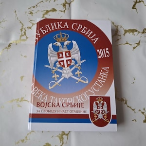 Può includere: Un libro con lo stemma serbo sulla copertina, raffigurante un'aquila bicipite, una corona e uno scudo con una croce. Il titolo include l'anno 2015 e testo in serbo. Il libro ha un bordo rosso e bianco.