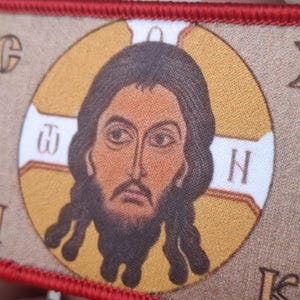 Peut inclure: Un &eacute;cusson rectangulaire avec une ic&ocirc;ne religieuse. L'image centrale est une repr&eacute;sentation d&eacute;taill&eacute;e d'un homme aux cheveux et &agrave; la barbe fonc&eacute;s, dans un cercle dor&eacute;. L'&eacute;cusson a une bordure rouge et comprend des lettres grecques.