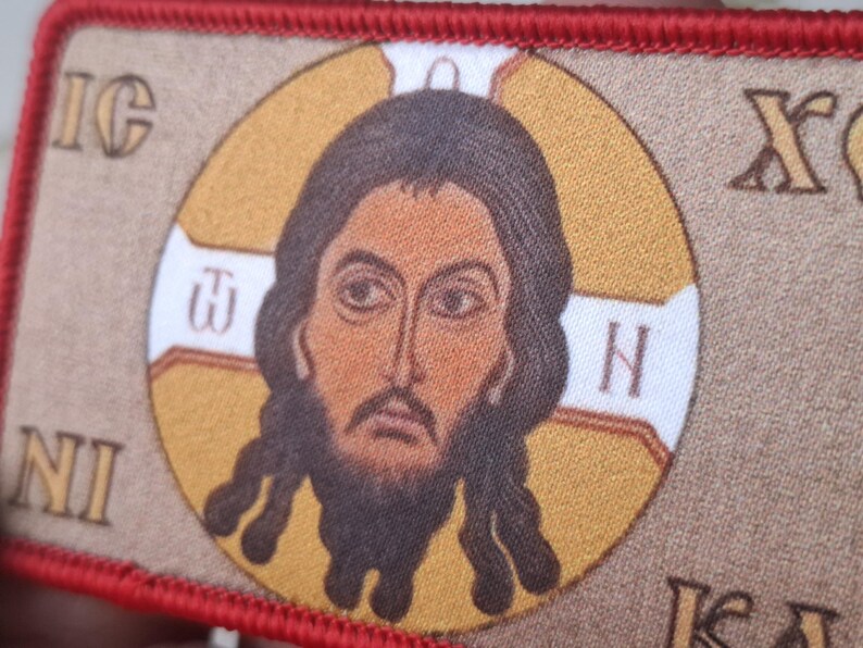 Peut inclure: Un &eacute;cusson rectangulaire avec une ic&ocirc;ne religieuse. L'image centrale est une repr&eacute;sentation d&eacute;taill&eacute;e d'un homme aux cheveux et &agrave; la barbe fonc&eacute;s, dans un cercle dor&eacute;. L'&eacute;cusson a une bordure rouge et comprend des lettres grecques.