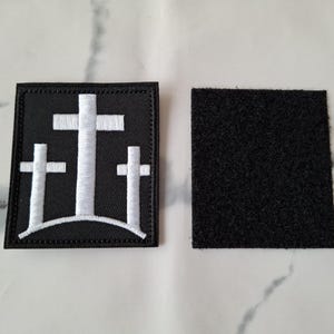 May include: A black square patch with three white crosses, one large in the center and two smaller ones on either side. A separate black square with a hook and loop backing is also shown.