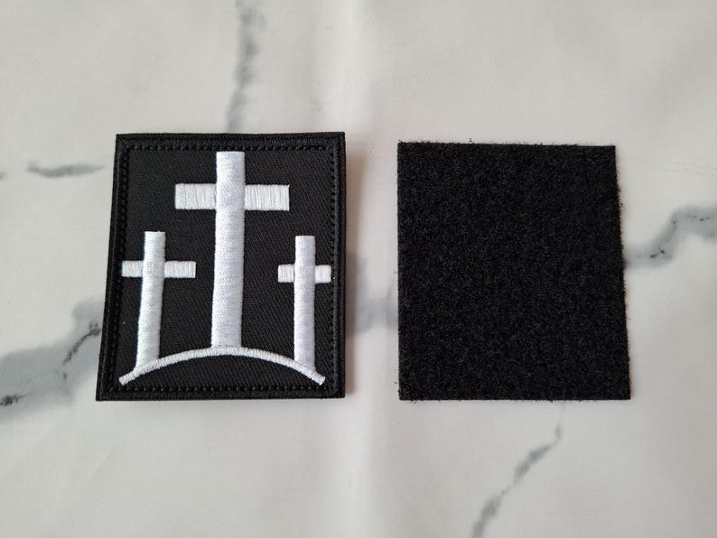 May include: A black square patch with three white crosses, one large in the center and two smaller ones on either side. A separate black square with a hook and loop backing is also shown.
