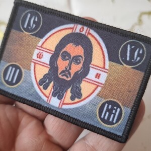 Peut inclure: &Eacute;cusson rectangulaire &agrave; bordure noire repr&eacute;sentant une ic&ocirc;ne religieuse. L'image centrale est une repr&eacute;sentation d'un homme avec une barbe et de longs cheveux, entour&eacute; d'un motif circulaire. L'&eacute;cusson comprend des lettres et des symboles grecs dans chaque coin, avec une palette de couleurs marron, or et bleu.