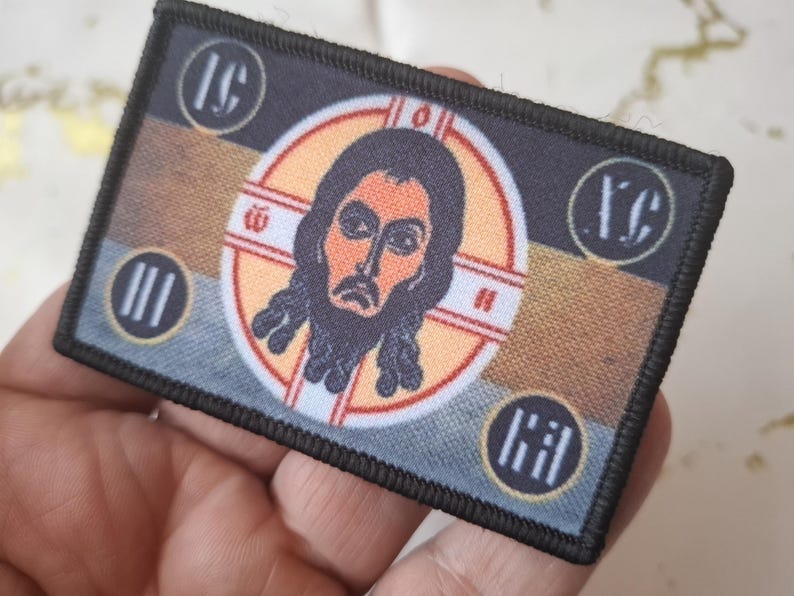 Peut inclure: &Eacute;cusson rectangulaire &agrave; bordure noire repr&eacute;sentant une ic&ocirc;ne religieuse. L'image centrale est une repr&eacute;sentation d'un homme avec une barbe et de longs cheveux, entour&eacute; d'un motif circulaire. L'&eacute;cusson comprend des lettres et des symboles grecs dans chaque coin, avec une palette de couleurs marron, or et bleu.